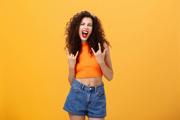 Femme berçant et profitant d'un grand concert dansant hurlant d'étonnement et de frisson montrant un geste rock-n-roll ouvrant la bouche et plissant posant dans une tenue élégante sur fond orange - Photo, image