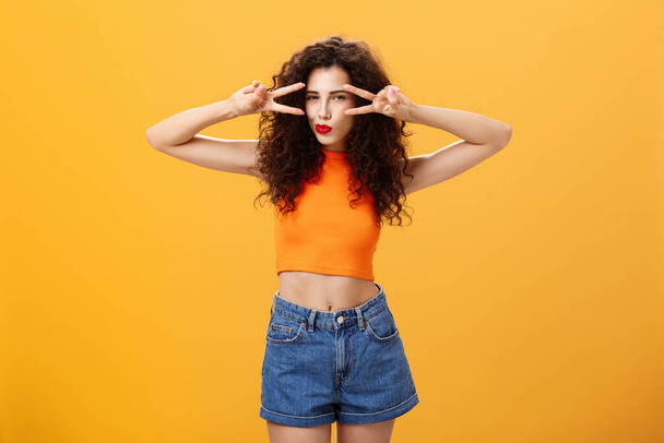 Portrait of good-looking daring and sexy stylish woman with curly hairstyle in red lipstick and cropped top showing victory or peace gestures overs eyes squinting folding lips flirty over orange wall - Foto, immagini
