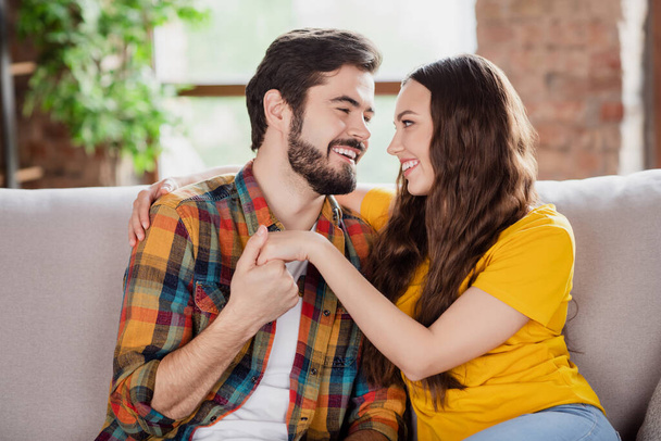 Photo of dreamy sweet young lovers dressed casual clothes sitting sofa smiling holding arms looking eyes indoors loft apartment - Φωτογραφία, εικόνα