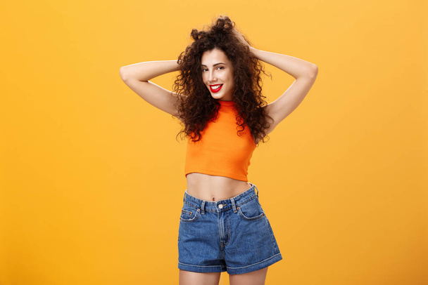 Woman posing for boyfriend being pleased with new look after visiting hairdresser touching curly hairstyle flirty and carefree smiling sensualy standing over orange background in cropped top - Photo, Image