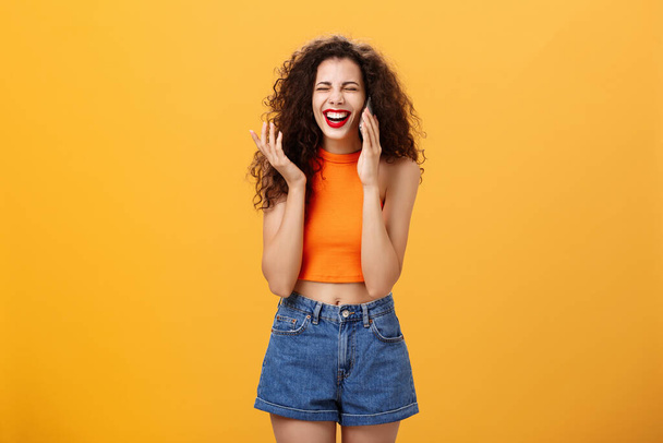 Общительный стильный и популярный европейский женщина с кудрявой прической и красной помадой смеется вслух, весело разговаривая на смартфоне с другом, играя с прядью волос беззаботно над оранжевой стеной - Фото, изображение
