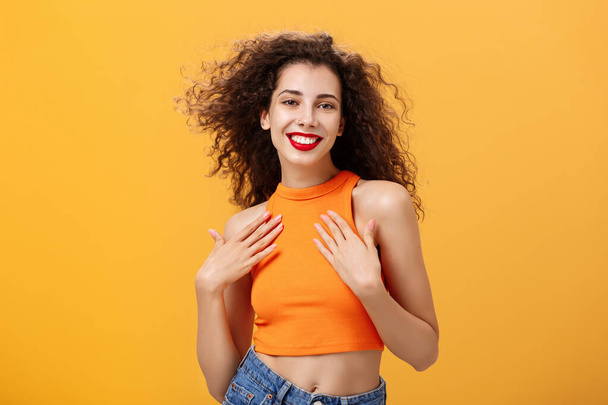 Waist-up shot of sensual and feminine tender female with curly hairstyle in cropped top holding palms on chest delighted and pleased smiling broadly posing flirty and sexy over orange wall - Photo, image