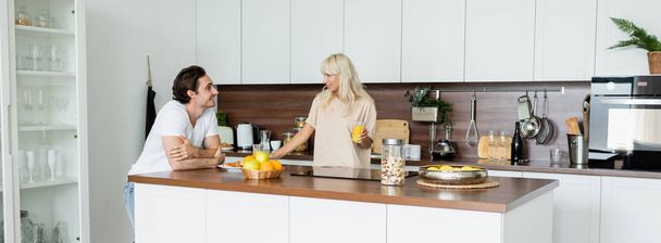 happy man looking at cheerful girlfriend with glass of orange juice in kitchen, banner - Photo, Image