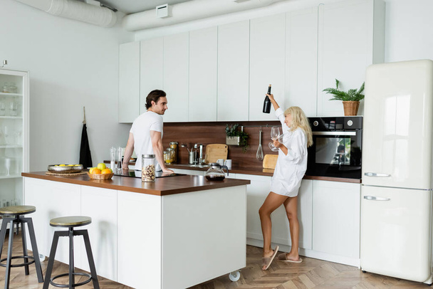 cheerful woman holding bottle of wine and glasses near boyfriend in kitchen  - Photo, Image