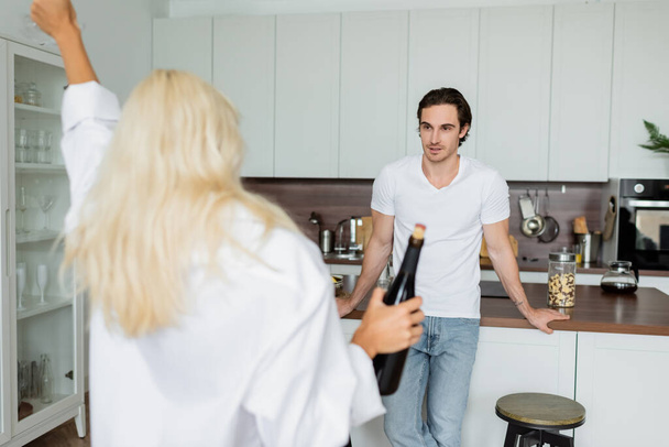 tattooed man looking at blurred blonde woman with bottle of wine in kitchen  - Foto, Imagen