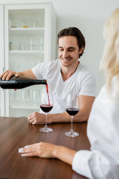 vidám férfi vörösbort önt az üvegbe a homályos barátnő közelében  - Fotó, kép