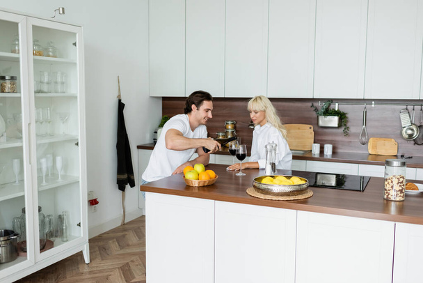 Счастливчик наливает красное вино в бокал рядом с блондинкой на кухне  - Фото, изображение
