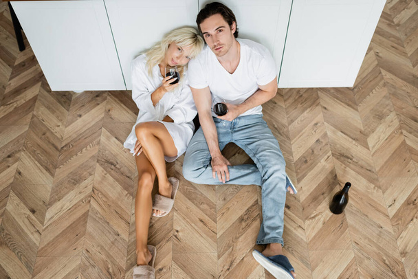 top view of young man and woman holding glasses of red wine while sitting on kitchen floor - Foto, Imagem