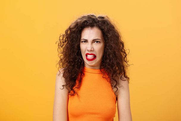Girl being disgusted with row food sticking out tongue and frowning showing aversion and disgust talking about thing she dislikes with friend standing intense and dissatisfied over orange background - Foto, afbeelding