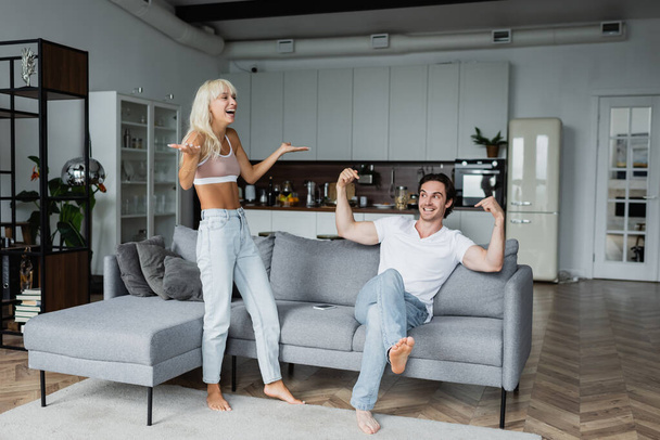 happy woman gesturing near cheerful boyfriend pointing at himself while sitting on sofa - Photo, Image