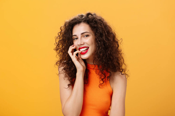Waist-up shot of charming silly and carefree feminine woman with curly hairstyle in cropped top biting finger and smiling sensually and flirty at camera looking sexy and daring over orange background - Foto, imagen