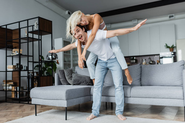 happy man with outstretched hands piggybacking blonde girlfriend in living room  - 写真・画像