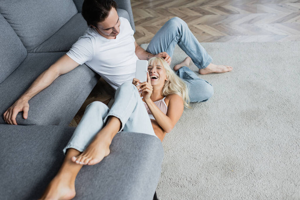 mujer alegre riendo y el uso de teléfono inteligente mientras se encuentra cerca de novio en la sala de estar  - Foto, imagen