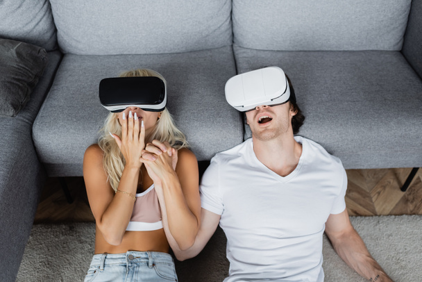 high angle view of shocked couple in vr headsets sitting near grey couch - Fotoğraf, Görsel