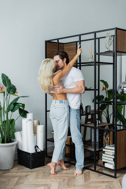 full length of blonde woman hugging man with closed eyes near rack in living room - Photo, Image