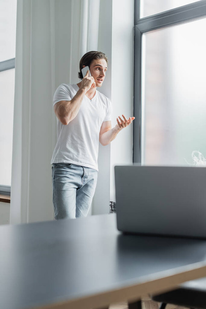 молодой человек жестикулирует во время разговора на смартфоне рядом с размытым ноутбуком на столе - Фото, изображение