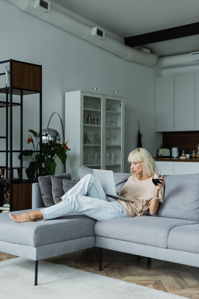 mujer rubia sosteniendo un vaso de vino tinto mientras usa el portátil en la sala de estar  - Foto, imagen