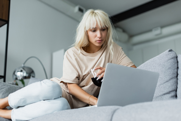 blonde freelancer holding glass of red wine while using laptop in living room  - Zdjęcie, obraz