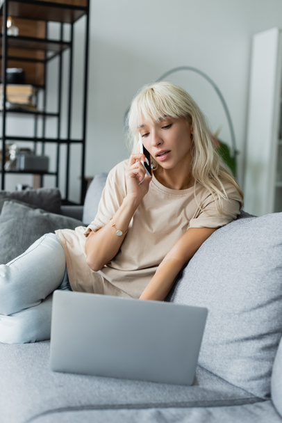 blonde freelancer talking on smartphone near blurred laptop on couch  - Zdjęcie, obraz