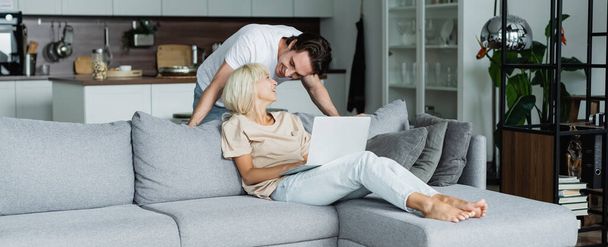 happy man looking at blonde girlfriend working from home with laptop, banner  - Foto, imagen