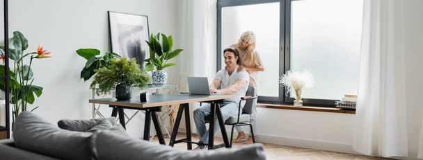 blonde woman massaging shoulders of boyfriend working from home with laptop, banner - Foto, Bild