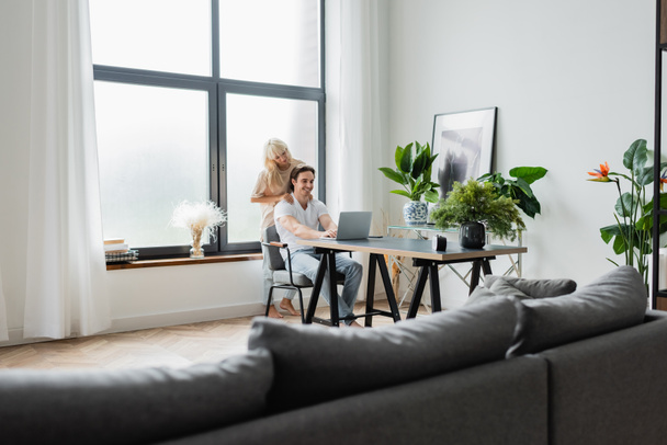 blonde woman massaging shoulders of boyfriend working from home with laptop - Фото, изображение