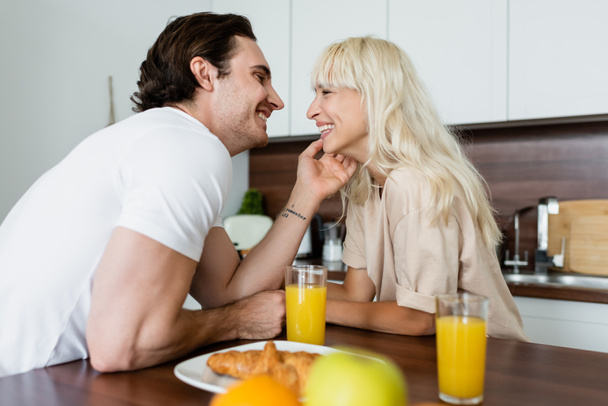 homme tatoué touchant visage de petite amie heureuse près de verres avec du jus d'orange et croissant - Photo, image