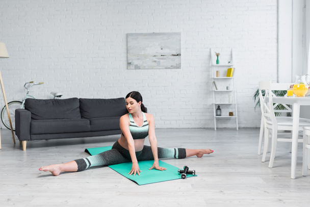 беременная женщина растягивается в сплит позе на коврике для йоги в гостиной - Фото, изображение