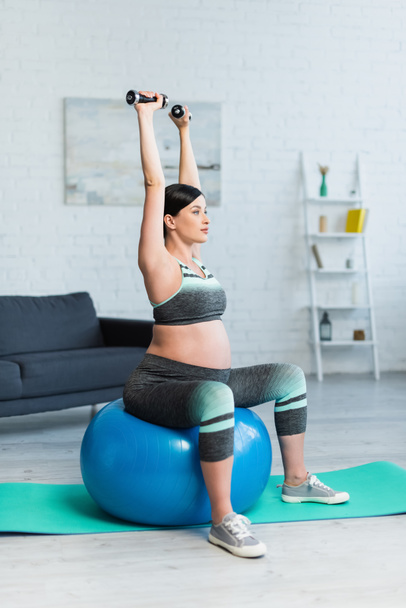 mujer embarazada joven sentada en la pelota de fitness y el entrenamiento con pesas en casa - Foto, Imagen