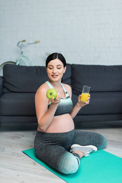 mujer embarazada feliz con manzana y jugo de naranja sentado con las piernas cruzadas en la alfombra de fitness - Foto, imagen