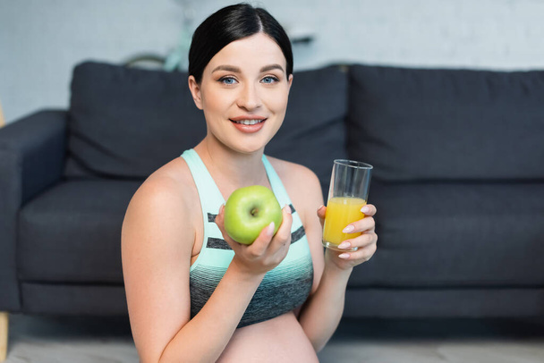 young pregnant woman in sports bra smiling at camera while holding apple and orange juice - Φωτογραφία, εικόνα
