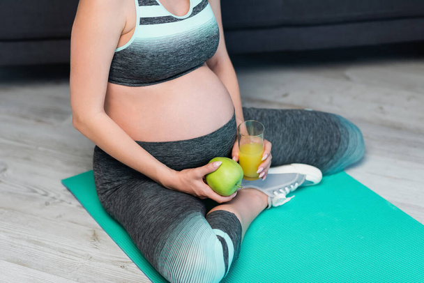 cropped view of pregnant woman in sportswear sitting on fitness mat with apple and orange juice - Valokuva, kuva