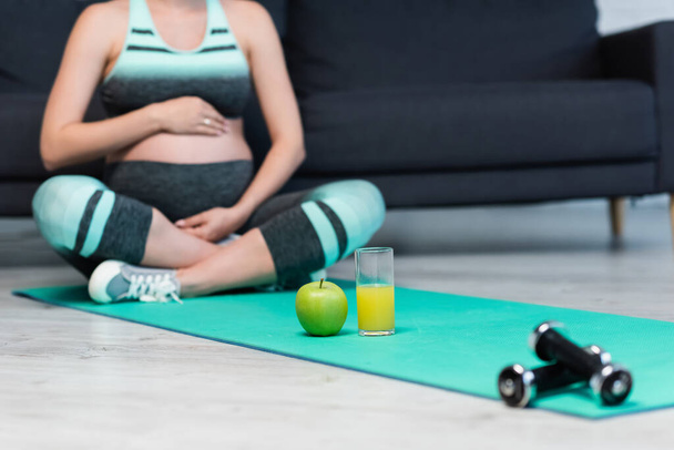 abgeschnittene Ansicht einer unscharfen Schwangeren, die in Lotuspose in der Nähe von frischem Apfel, Orangensaft und Hanteln meditiert - Foto, Bild