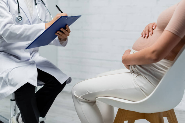 vista recortada de la mujer embarazada abrazando el vientre mientras está sentado cerca del médico escribiendo en el portapapeles - Foto, Imagen