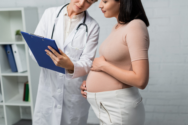 médico mostrando la prescripción en portapapeles a la mujer embarazada en la sala de consulta - Foto, Imagen