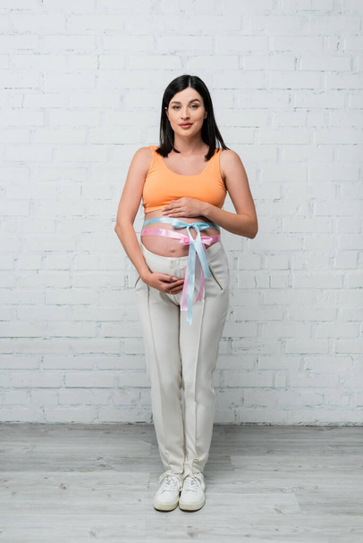 junge schwangere Frau steht an weißer Wand und berührt Bauch mit Satinbändern  - Foto, Bild