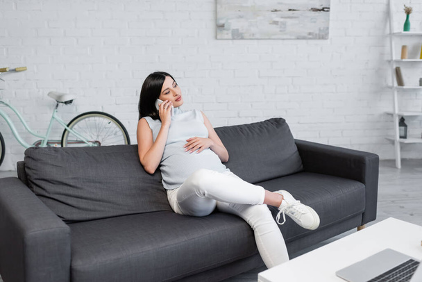 young pregnant woman talking on smartphone in living room near blurred laptop on table - Photo, Image