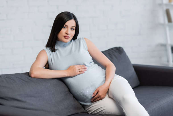 young pregnant woman embracing belly while sitting on couch at home - Fotografie, Obrázek