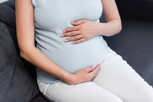 vista parcial de la mujer embarazada abrazando el vientre mientras está sentada en el sofá - Foto, imagen