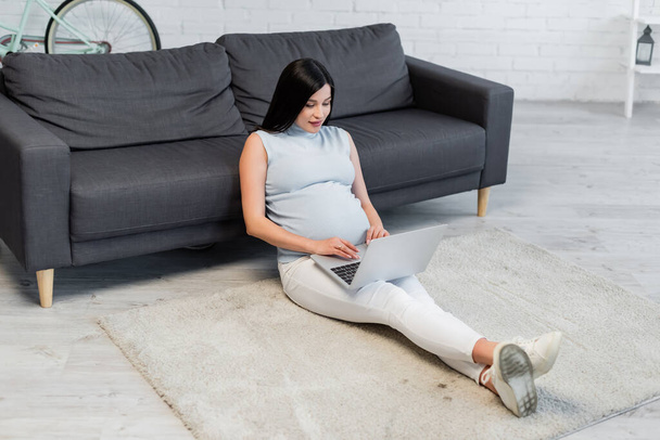 young pregnant woman sitting on floor near couch and typing on laptop - Фото, изображение