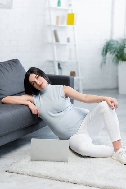 smiling pregnant woman looking at camera while sitting on floor near laptop and sofa - Foto, immagini