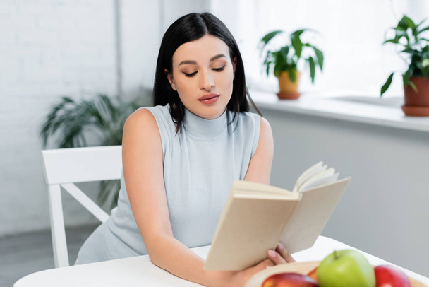 junge Frau liest Buch neben verschwommenen Äpfeln auf Küchentisch - Foto, Bild