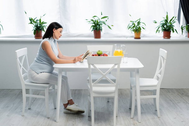 side view of pregnant woman reading book near orange juice and fresh apples on table in kitchen - Photo, Image