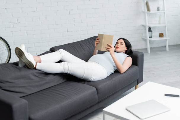 terhes nő olvasás könyvet, miközben feküdt a kanapén közelében elmosódott kütyü az asztalon a nappaliban - Fotó, kép