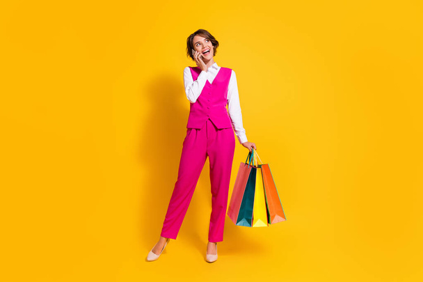 Photo of pretty dreamy young lady wear pink suit talking modern device holding bargains looking empty space isolated yellow color background - Fotó, kép