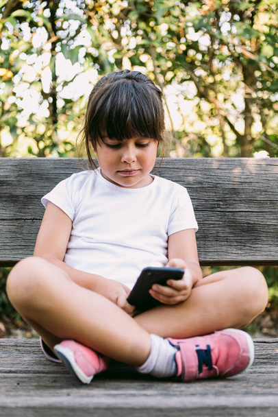 Little black-haired girl, wearing a white t-shirt, sitting on a bench in a park, looking at the mobile phone, smiling. - Φωτογραφία, εικόνα