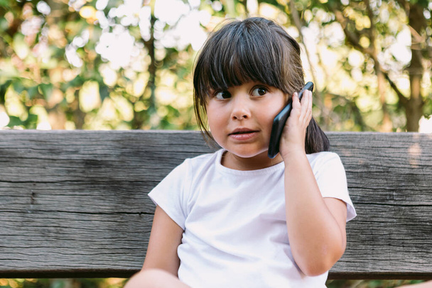 Little girl with black hair, wearing a white t-shirt, sitting on a bench in a park talking on the mobile phone, with a serious gesture. - Foto, Imagen