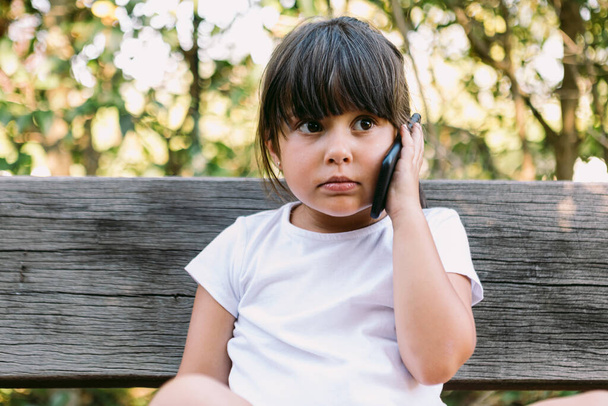 Little girl with black hair, wearing a white t-shirt, sitting on a bench in a park talking on the mobile phone, with a serious gesture. - Φωτογραφία, εικόνα