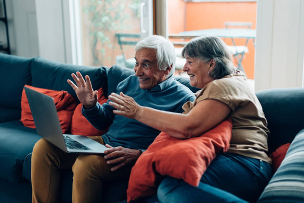 pareja de ancianos saluda durante una videollamada - personas mayores en el hogar conectado inalámbrico a Internet mediante la aplicación de llamada en el ordenador portátil - Foto, Imagen