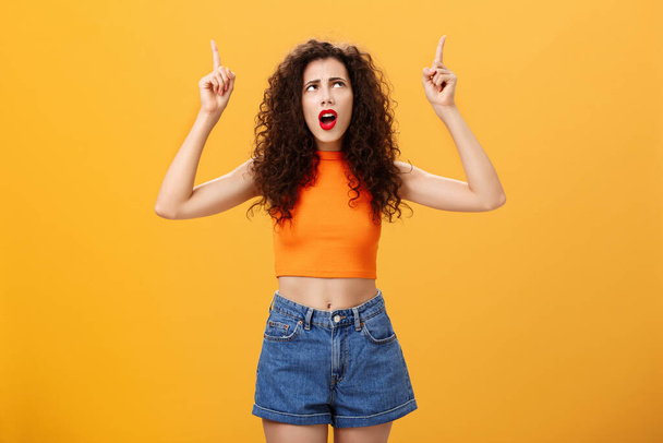 Silly and moody european party girl with curly hairstyle in red lipstic and stylish orange cropped top complaining on bad weather raising hands pointing and looking up with displeased unhappy face - Photo, Image